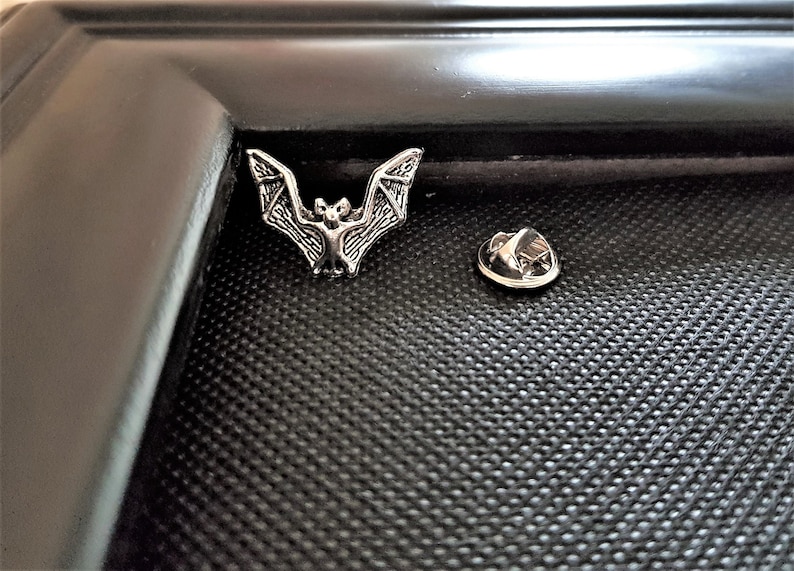 May include: A silver-toned bat pin with a detailed design. The pin is on a black background and includes a separate pin back.