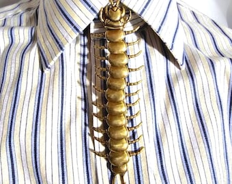 Könnte beinhalten: Eine goldfarbene Tuchklemme in Form eines Hundertfüßers, getragen auf einem gestreiften Button-Down-Hemd. Das insektenförmige Accessoire hat ein detailliertes Design mit segmentiertem Körper und Beinen. Die Krawattenklammer verleiht dem Hemd eine einzigartige Note.