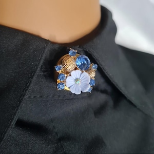 May include: A gold-toned brooch featuring a light blue flower with a central iridescent gem, surrounded by blue gemstones and textured gold accents. The brooch is attached to a black shirt.