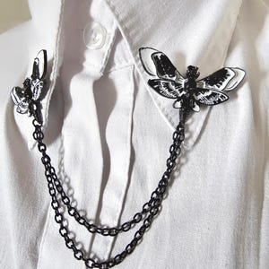K&ouml;nnte beinhalten: Eine schwarz-wei&szlig;e Mottenbrosche mit Kette, befestigt an einem wei&szlig;en Hemd mit Kragen. Die Brosche hat zwei Mottenmotive, eines auf jeder Seite des Kragens, die durch eine Doppelkette verbunden sind. Die Motten haben detaillierte Fl&uuml;gelmuster.