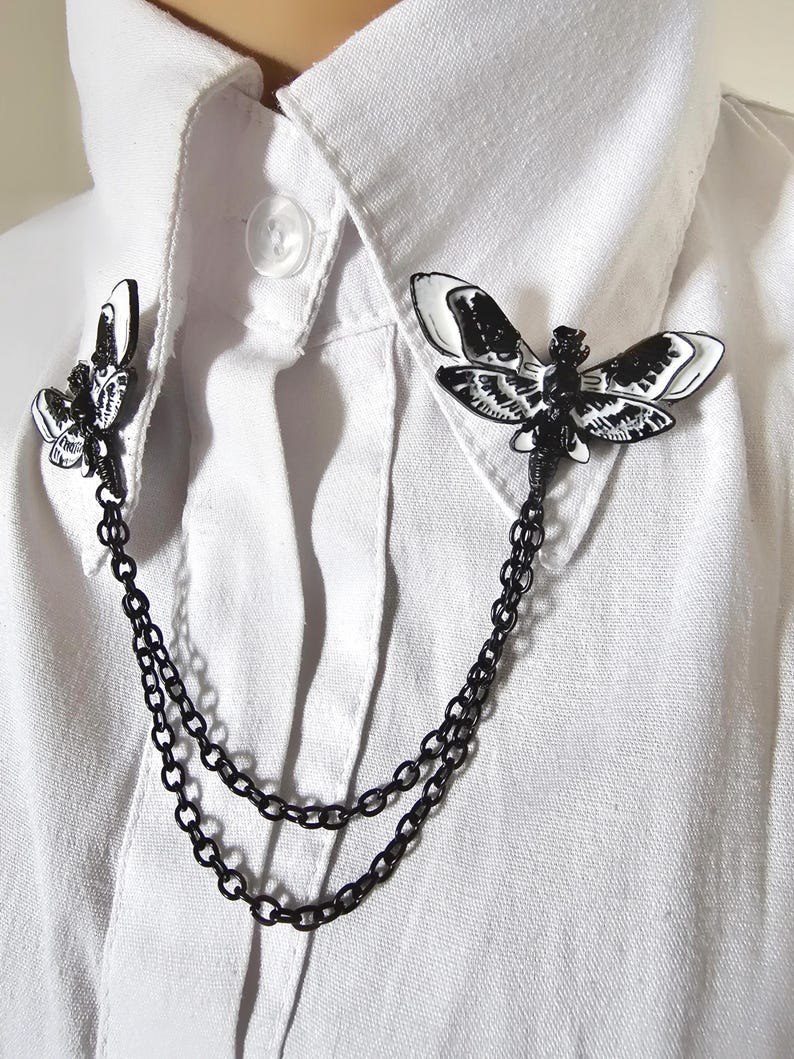 K&ouml;nnte beinhalten: Eine schwarz-wei&szlig;e Mottenbrosche mit Kette, befestigt an einem wei&szlig;en Hemd mit Kragen. Die Brosche hat zwei Mottenmotive, eines auf jeder Seite des Kragens, die durch eine Doppelkette verbunden sind. Die Motten haben detaillierte Fl&uuml;gelmuster.