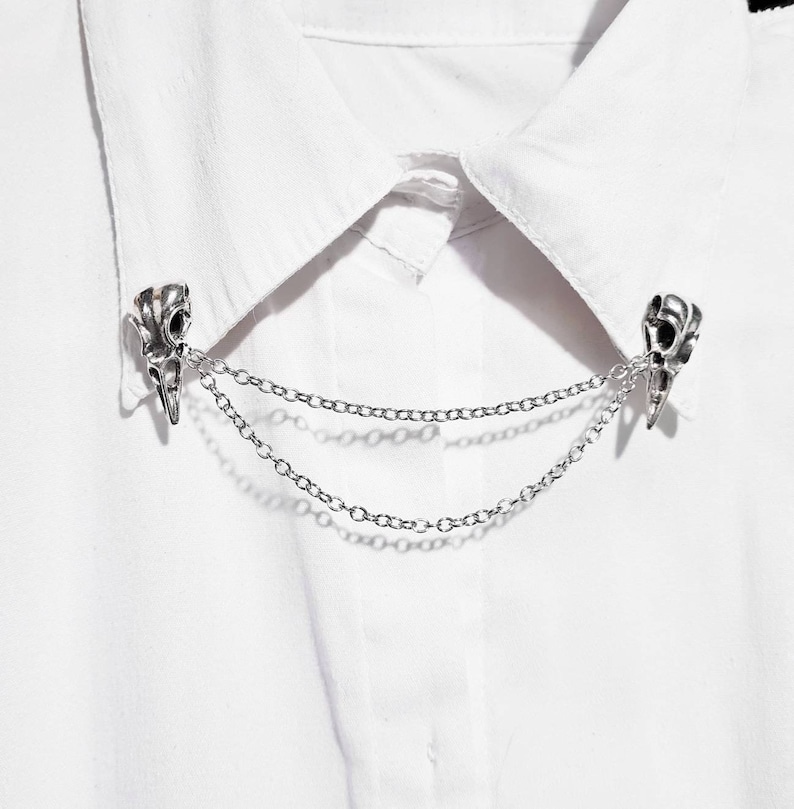 Puede incluir: Pin de collar plateado con dos abalorios en forma de calavera conectados por una cadena doble.