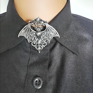 Könnte beinhalten: Silberfarbene Fledermaus-Brosche mit aufwendigen Filigranflügeln und -körper, auf einem schwarzen Hemd mit Kragen. Die Brosche hat ein detailliertes Design, perfekt, um jedem Outfit einen gotischen Touch zu verleihen.