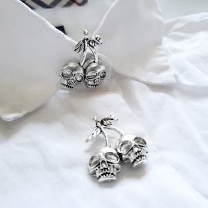 May include: Silver-toned skull-shaped cherry collar pins on a white collared shirt. The pins feature two skulls connected by a stem and leaves, adding a unique, gothic touch to the shirt.