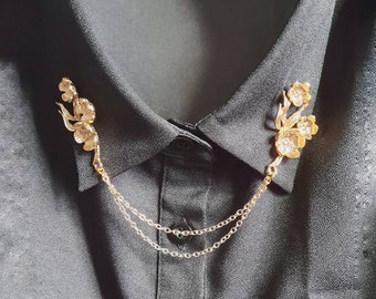 Può includere: Un fermaglio per colletto dorato con un design floreale e una doppia catena. Il fermaglio è attaccato a una camicia nera.