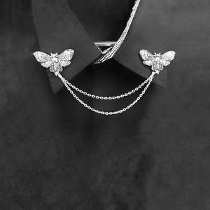 May include: A silver-toned collar pin set featuring two moth-shaped brooches with intricate wing patterns and a central skull detail. The two moth pieces are connected by two delicate silver-toned chains that drape across the front of the collar.