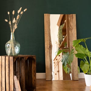 Peut inclure: Un grand miroir rectangulaire avec un cadre en bois naturel est posé sur un sol en bois. Le miroir reflète une pièce avec des meubles en bois et des plantes. Un vase en verre transparent avec des plantes séchées est posé sur une caisse en bois.