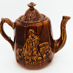 May include: A brown ceramic teapot with a lid and a handle. The teapot has a raised design of a person standing next to a barrel. The text "BE STILL TO KNOW" is on the bottom of the teapot.