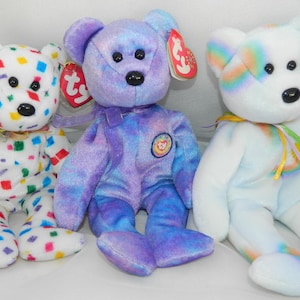 May include: Three plush teddy bears with black plastic eyes and noses. The left bear is white with multicolored square confetti patterns. The center bear is purple with a tie-dye effect and a circular patch. The right bear is white with rainbow accents.