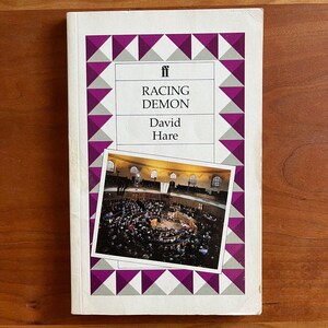 May include: A paperback book titled "RACING DEMON" by David Hare. The cover features a photograph of a theater interior with an audience. The title is in a white rectangle with a purple and white geometric border. The publisher's logo is at the top.