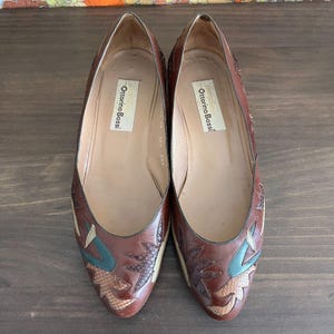May include: A pair of brown leather Ottorino Bossi flats. The shoes feature decorative accents in shades of green, tan, and snakeskin-like patterns. The interior of the shoes is a light tan color, and the brand name is visible on the insole.