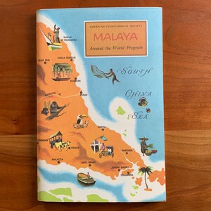 May include: A vintage travel brochure titled "Malaya" from the American Geographical Society. The cover features a colourful illustrated map of the region, with text and illustrations of local scenes and landmarks. The brochure is open on a wooden surface.