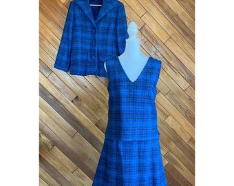 May include: A vintage blue plaid dress and matching jacket set. The sleeveless dress has a V-neck and a flared skirt. The jacket has a notched collar and button closure. The set is displayed on a mannequin against a wooden background.