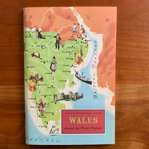 Op de afbeelding: Een vintage kaart van Wales uit het "Around the World Program" van de American Geographical Society. De kaart bevat illustraties van lokale bezienswaardigheden, mensen en dieren, met het Bristol Channel aan de rechterkant. De omslag ligt op een houten oppervlak.