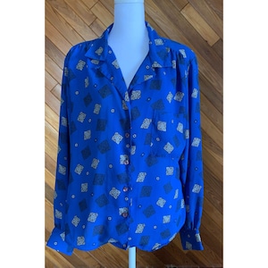 Puede incluir: Una camisa azul de manga larga con cuello y cierre de botones. La camisa presenta un estampado de formas geométricas negras y blancas. La camisa se exhibe en un maniquí blanco.