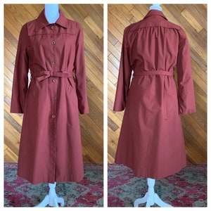 Könnte beinhalten: Ein langärmliger, burgunderfarbener Trenchcoat mit Knopfleiste und Bindegürtel. Der Mantel hat eine klassische A-Linien-Silhouette.