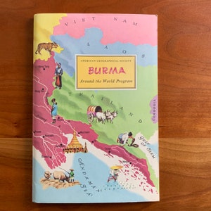 Op de afbeelding: Een vintage boekomslag getiteld "Burma" van de "Around the World Program" van de American Geographical Society. De omslag toont een kleurrijke kaart van Birma en de omliggende landen, met illustraties van mensen, dieren en bezienswaardigheden. Het kleurenpalet omvat roze, groen, geel en blauw.