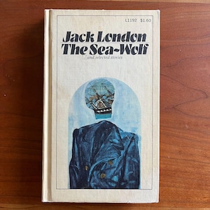 May include: Vintage paperback book titled "Jack London The Sea-Wolf and selected stories." The cover features a blue illustration of a ship and a blue coat. The book has a cream-colored spine and cover.