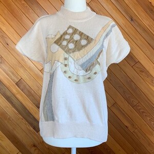 Puede incluir: Un suéter de color crema de manga corta con un diseño geométrico en tonos marrones, beige y grises.