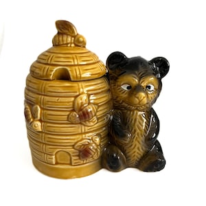 May include: A ceramic honey pot shaped like a beehive with a brown bear figurine next to it. The beehive has a lid with a bee on top and is decorated with bees and honeycomb patterns. The bear is brown with black eyes and a black nose.
