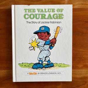 May include: A hardback book titled "The Value of Courage" with the subtitle "The Story of Jackie Robinson." The cover shows a baseball player in a blue uniform, holding a bat and a baseball. The book is a ValueTale by Spencer Johnson, M.D.