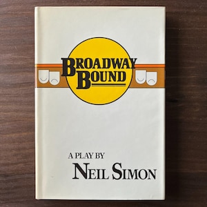May include: A vintage paperback book titled "Broadway Bound" by Neil Simon. The cover features a yellow circle with the title in bold black letters, and a brown and orange horizontal stripe with theatre masks. The background is off-white.
