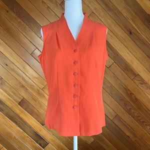 May include: A sleeveless, orange blouse with a V-neck and a row of six matching buttons down the front. The blouse is made of a smooth fabric and has a tailored fit, suitable for various occasions.
