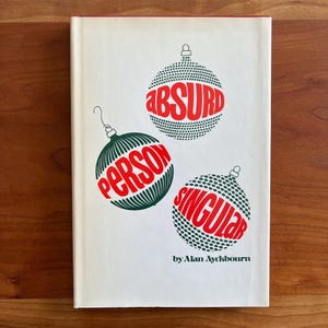 Op de afbeelding: Een vintage boekomslag met een witte achtergrond en rode rand. De omslag heeft drie groene en witte gestippelde ornamentontwerpen, elk met een rood woord: "ABSURD", "PERSON" en "SINGULAR". De naam van de auteur, Alan Ayckbourn, staat onderaan gedrukt.