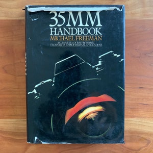 Op de afbeelding: Een vintage boek getiteld "35MM Handbook" van Michael Freeman. De omslag is zwart met witte en oranje afbeeldingen van een cameralens en andere cameraonderdelen. Het boek ligt op een houten oppervlak.