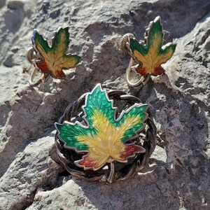 May include: A set of vintage jewelry featuring a brooch and a pair of earrings. The pieces are made of silver and feature a green, yellow, and red enamel maple leaf design.