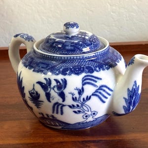 May include: A white ceramic teapot featuring a blue willow pattern. The design includes illustrations of two birds, a boat, and a tree. The teapot has a curved handle, a spout, and a matching lid with a small knob on top. The surface is glossy.