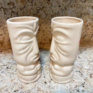 May include: Two off-white ceramic tiki mugs featuring a sculpted face with closed eyes, a prominent nose, and a small mouth. The mugs have a cylindrical shape with a smooth, glossy finish and a base that mimics a stylized torso and folded hands.