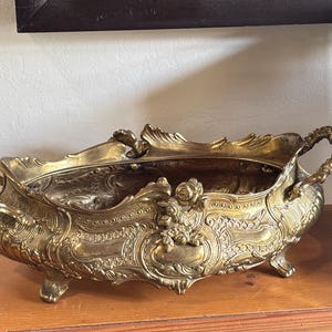 May include: An oval brass centerpiece bowl featuring ornate embossed scrollwork, floral motifs, and two decorative handles on the sides. The vessel is supported by four small feet and has a polished metallic finish with intricate relief detailing.