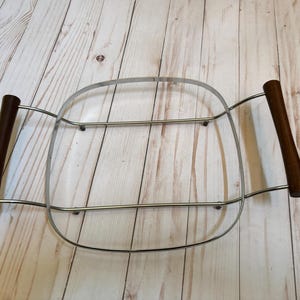 May include: A vintage metal serving tray with a square shape and a chrome finish. It features two dark brown wooden handles on each side. The tray is designed to hold a dish or bowl.