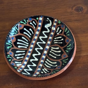 May include: A small, round, ceramic dish with a hand-painted design. The dish features a black background with brown, white, blue, and green floral and geometric patterns. The rim is a reddish-brown color.