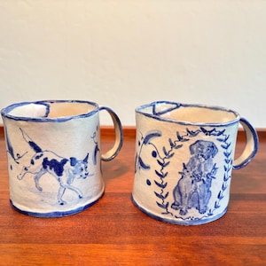 May include: Two handmade ceramic mugs with an off-white base and blue hand-painted illustrations. One mug features a dog running, while the other displays a seated dog framed by a leafy wreath. Both mugs have irregular, organic rims and handles.