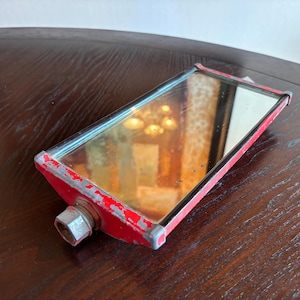 May include: Vintage red and silver side mirror with a rectangular shape. The mirror has a silver frame and a red metal casing with signs of wear. A hexagonal bolt is attached to the back.