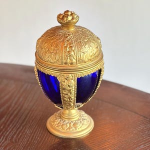 May include: An antique, ornate egg-shaped container with a gold-colored metal exterior and a deep blue glass interior. The lid and base feature intricate relief patterns. The container sits on a round, decorative base.