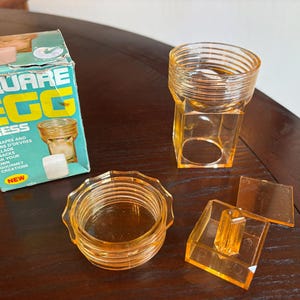 May include: A vintage Square Egg Press kitchen tool with a teal and yellow cardboard box. The set includes a transparent amber-colored plastic cylindrical screw-top lid, a square-shaped base container, and a matching square-shaped press insert.
