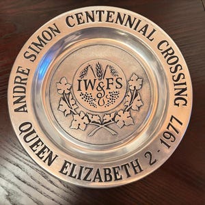 May include: A silver-colored plate with embossed text and a central emblem. The text reads "Andre Simon Centennial Crossing 1977" and "Queen Elizabeth 2". The emblem features wheat, grapes, and the letters "IW&FS".