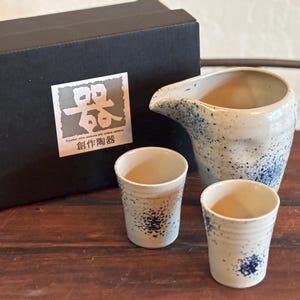 May include: A ceramic sake set with a pitcher and two cups, featuring a white and blue speckled design. The set is accompanied by a black box with Japanese text and a logo.