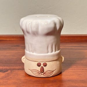 May include: A ceramic salt shaker shaped like a chef. The shaker is off-white with a chef's hat and a face with a brown mustache and nose. The salt shaker has small handles on each side of the face.