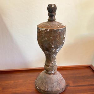 May include: A tall, turned wooden finial with a weathered brown finish and patches of white paint. The object features a rounded base, a slender central column, and a bulbous upper section topped with a small, spherical decorative knob.