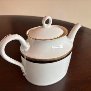 May include: White ceramic teapot with a curved handle and spout. The lid and rim are accented with gold trim, and a black band encircles the body. The teapot has a loop handle on the lid.