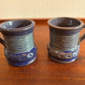 May include: Two small ceramic mugs with a glossy finish in shades of deep blue and muted purple. Each mug features a cylindrical body with horizontal ribbed detailing around the center and a decorative band of circular indentations near the base.