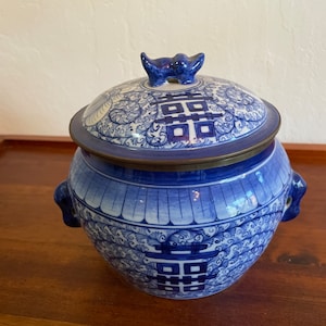 May include: A blue and white porcelain jar with a lid. The jar has a traditional Chinese design with a double happiness symbol. The lid has a small knob in the shape of a bird.