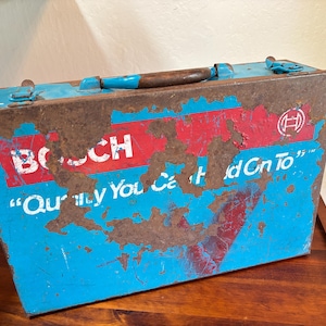 May include: A rectangular blue metal tool box with a brown handle and metal latches. The front features a red horizontal stripe with the white text BOSCH and the slogan "Quality You Can Hold On To" in white, with significant rust and paint wear.