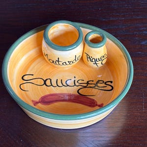 May include: A ceramic serving dish with a yellow and green rim, featuring two small condiment pots and the word "Saucisses." The dish has a painted sausage graphic. The pots are labeled "Moutarde" and "Pique."