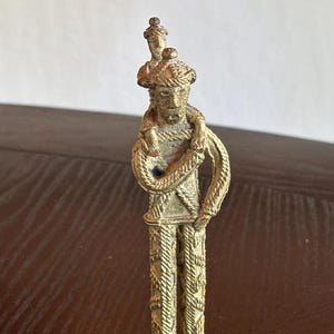 May include: A small, antique brass figurine depicting a person carrying another person on their shoulders. The figures are adorned with detailed clothing and headwear. The figurine has a weathered appearance, suggesting age and use.