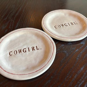 May include: Two off-white ceramic plates with the word "COWGIRL" embossed in the center. The plates have a dotted red border. The plates are on a dark brown wooden surface.
